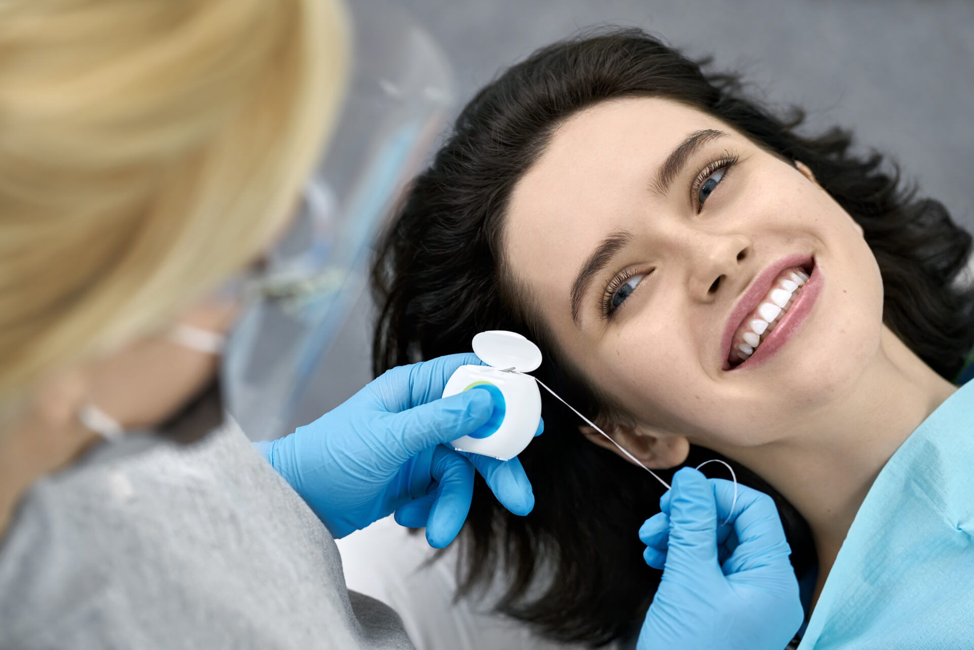 Glad girl in a patient's bib in a dental clinic. Dentist in blue latex gloves prepares to flossing her teeth with a help of a dental floss. Closeup. Horizontal.